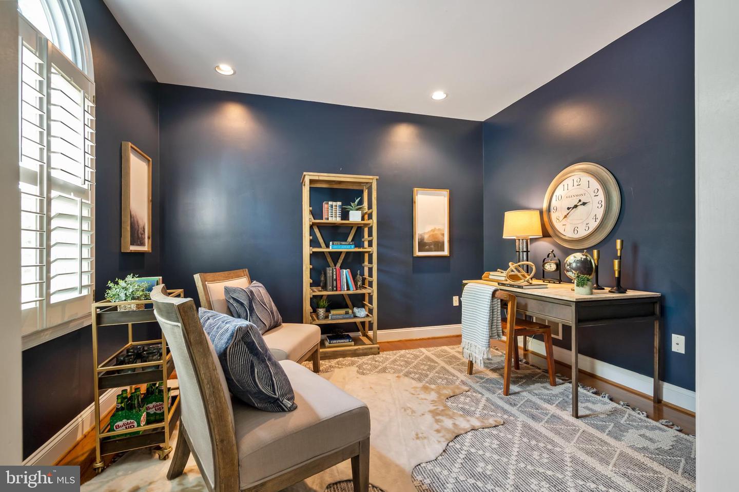 Staged home office with navy walls, plantation shutters, and a wooden desk and bookcase.