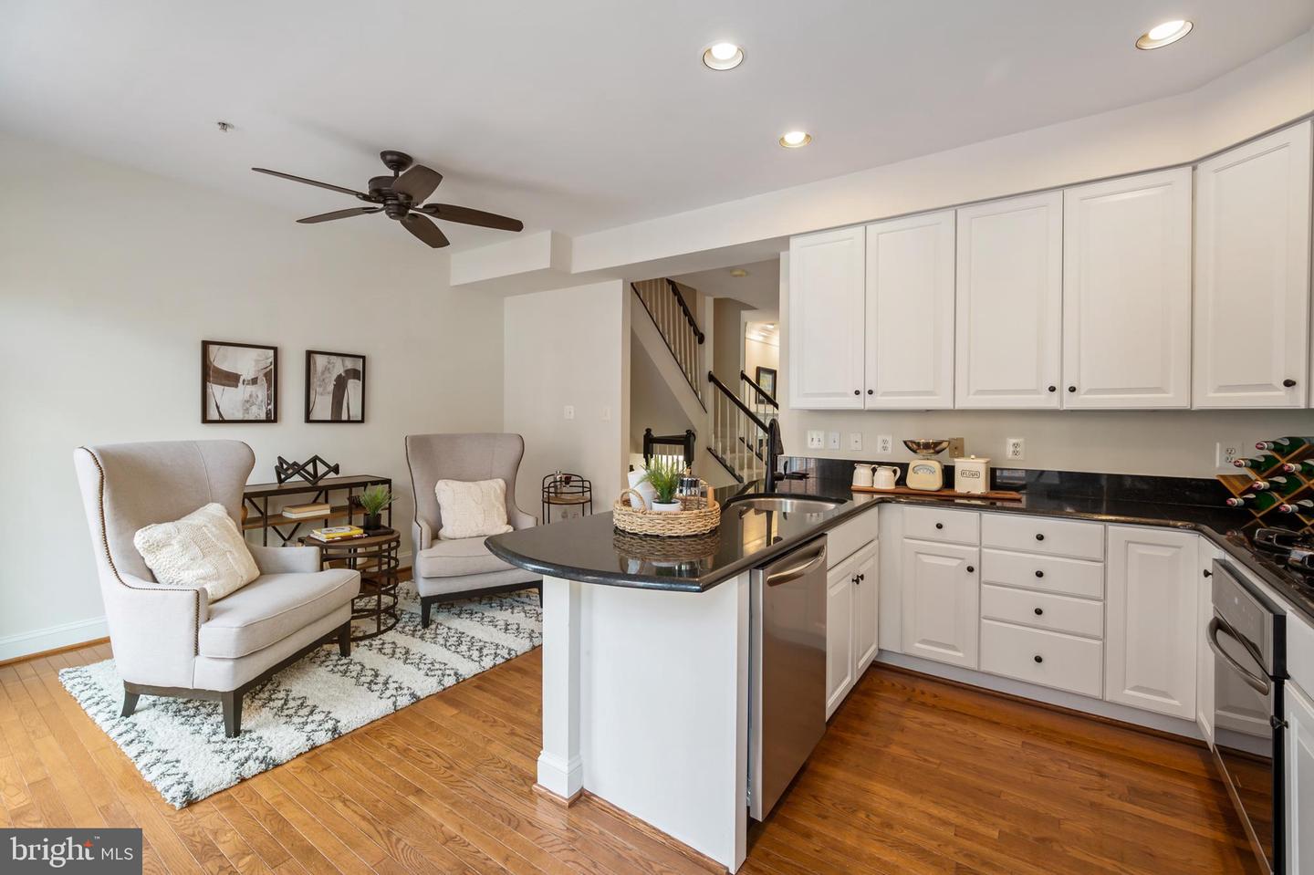 Open kitchen and sitting area with white cabinets, a dark stone peninsula, hardwood floors, and upholstered chairs.