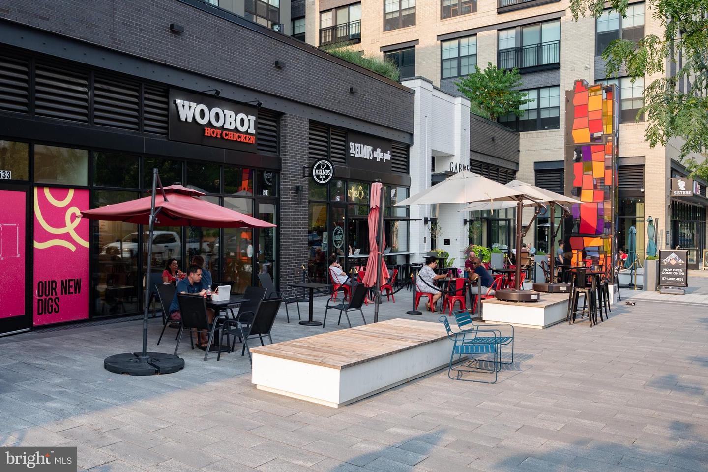 Pedestrian plaza with restaurant patios, colorful art installation, and modern apartment buildings.