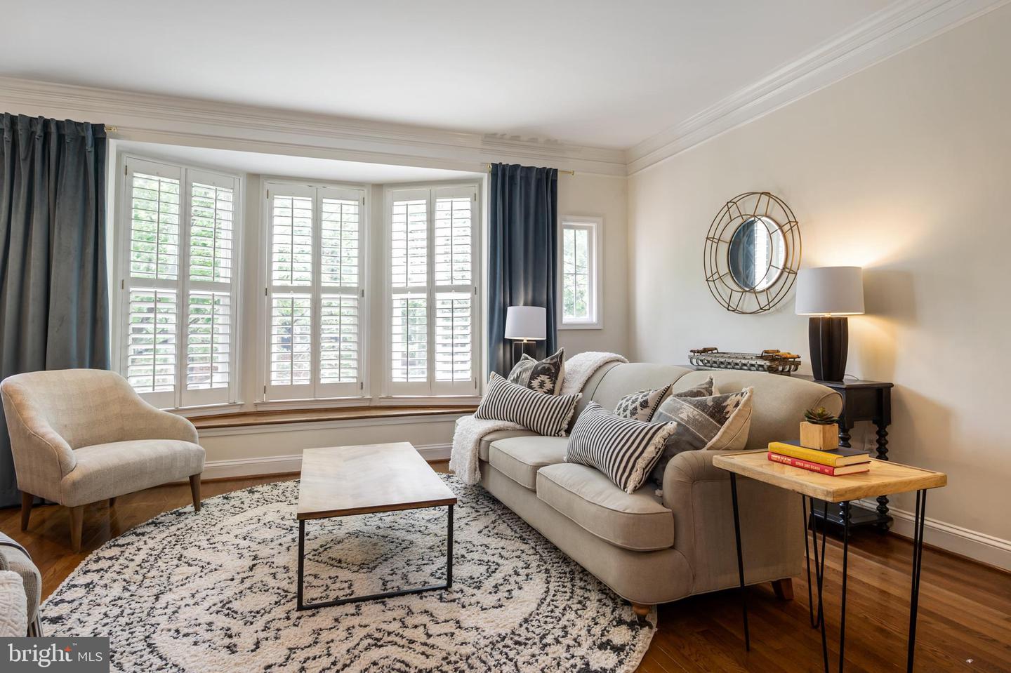 Living room with a shuttered bay window, beige seating, patterned rug, and hardwood floors.