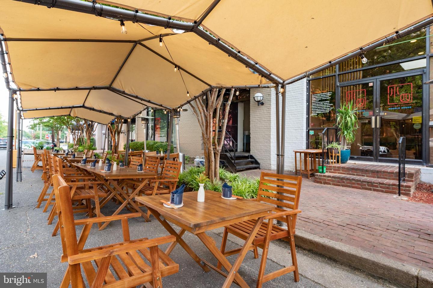 Restaurant patio with wooden tables beneath tan awnings beside a glass-fronted noodle house.