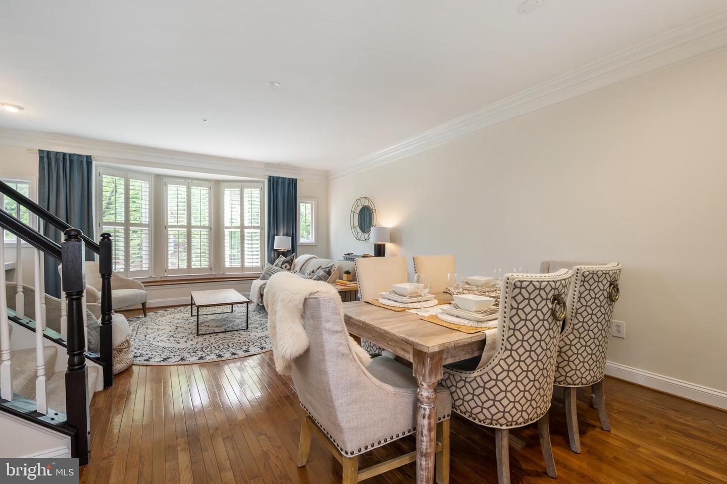 Staged living and dining area with hardwood floors, shuttered windows, and a dark-railed staircase.