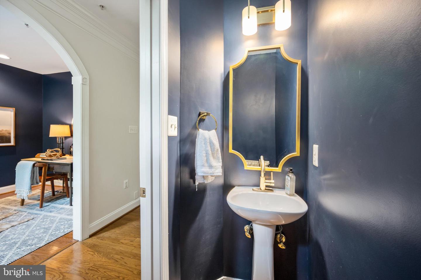 Powder room with a white pedestal sink, gold-framed mirror, and dark navy walls beside an arched doorway.