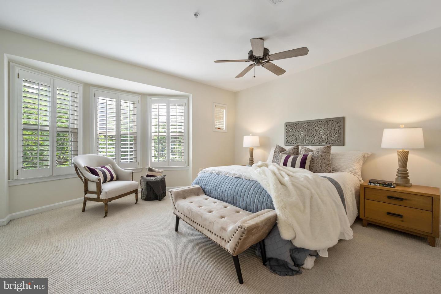 Staged bedroom with a tufted bench, ceiling fan, and wide plantation-shuttered windows.