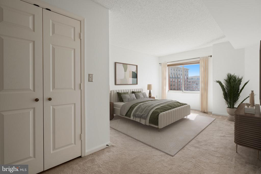 Furnished bedroom with gray and green bedding, abstract art, and a window overlooking nearby buildings.