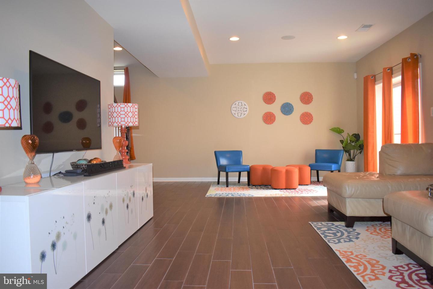Furnished living room with a wall-mounted television, beige sectional, blue chairs, and orange ottomans.