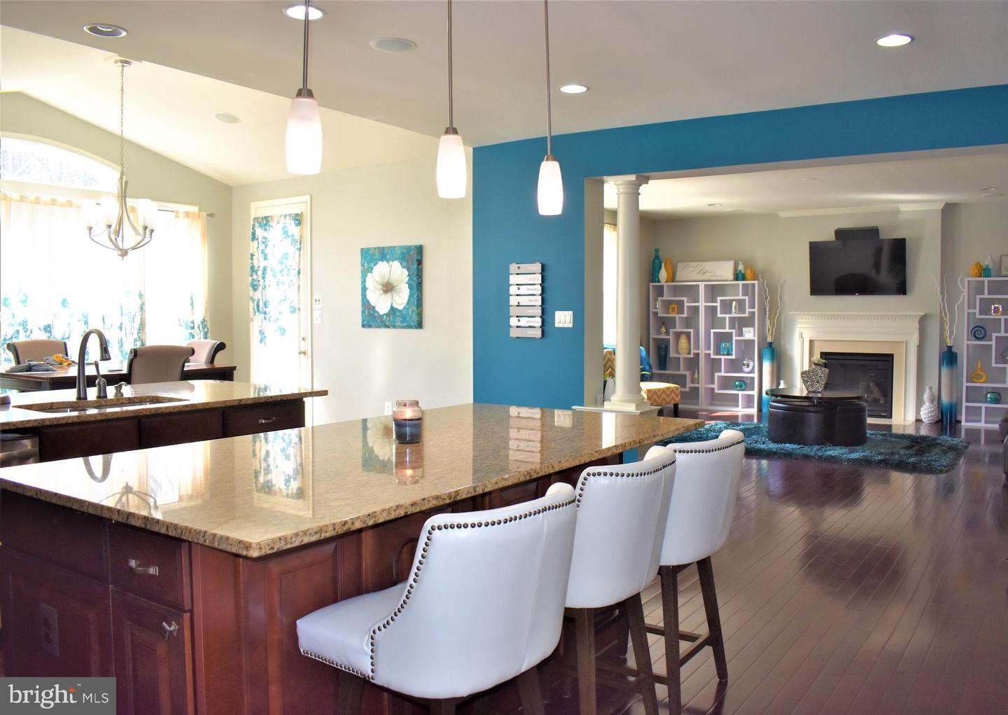 Open kitchen with a granite island and white stools adjoining a living room with a fireplace.