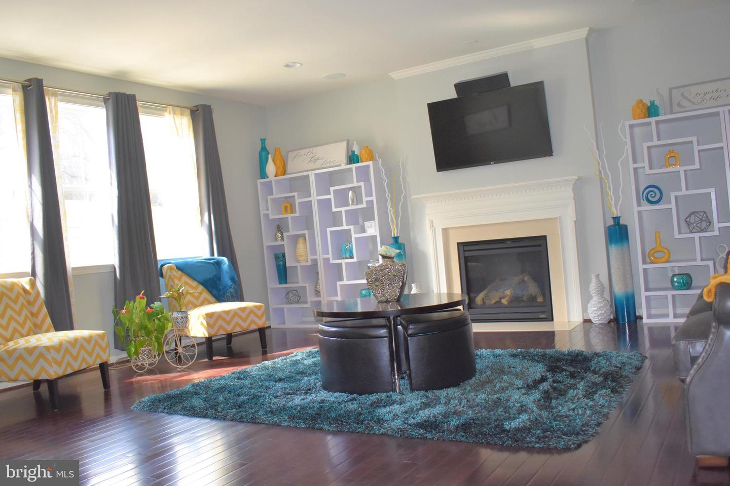Staged living room with a white fireplace, mounted TV, turquoise rug, and tall curtained windows.