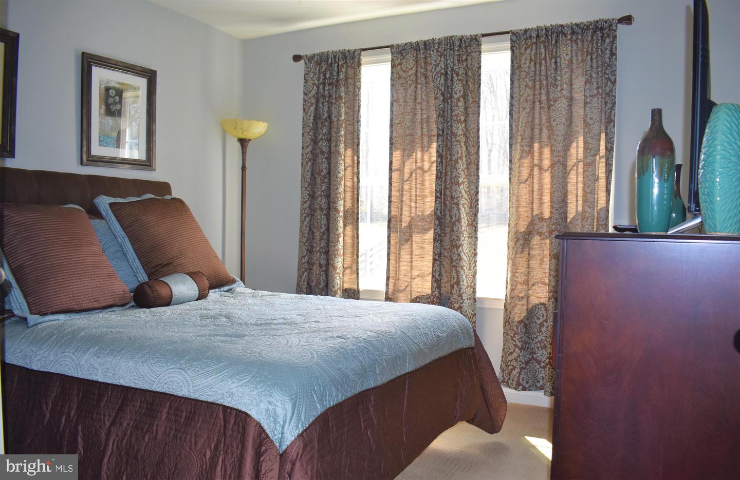 Furnished bedroom with a blue-and-brown bedspread, patterned curtains, and a wooden dresser with vases.