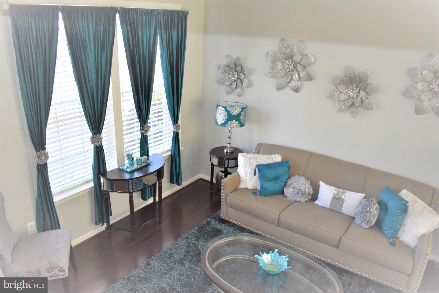Staged living room with a beige sofa, teal curtains, silver floral wall decor, and dark wood floors.