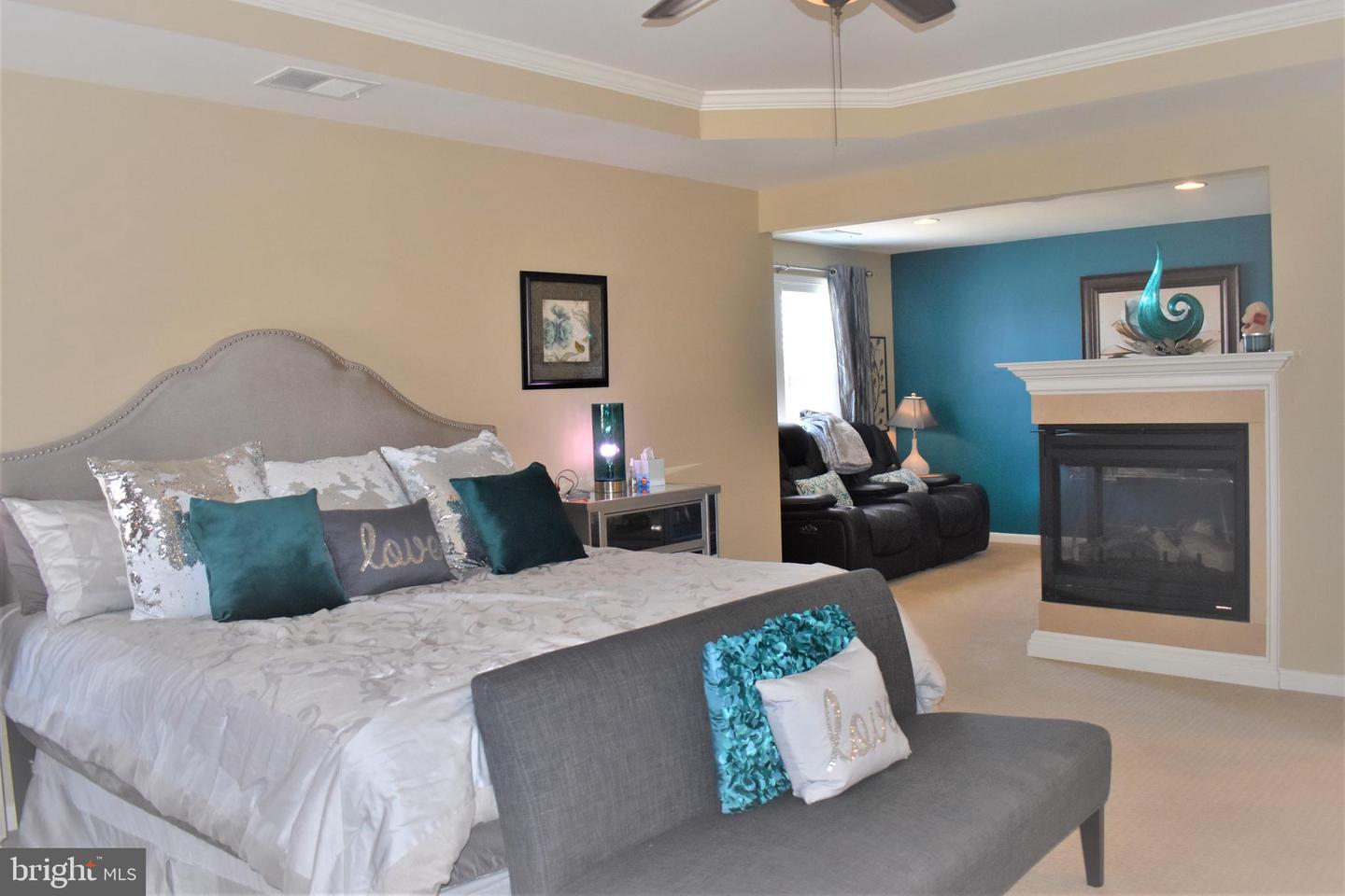 Staged bedroom with an upholstered bed, gray chaise lounge, and fireplace beside a teal sitting area.