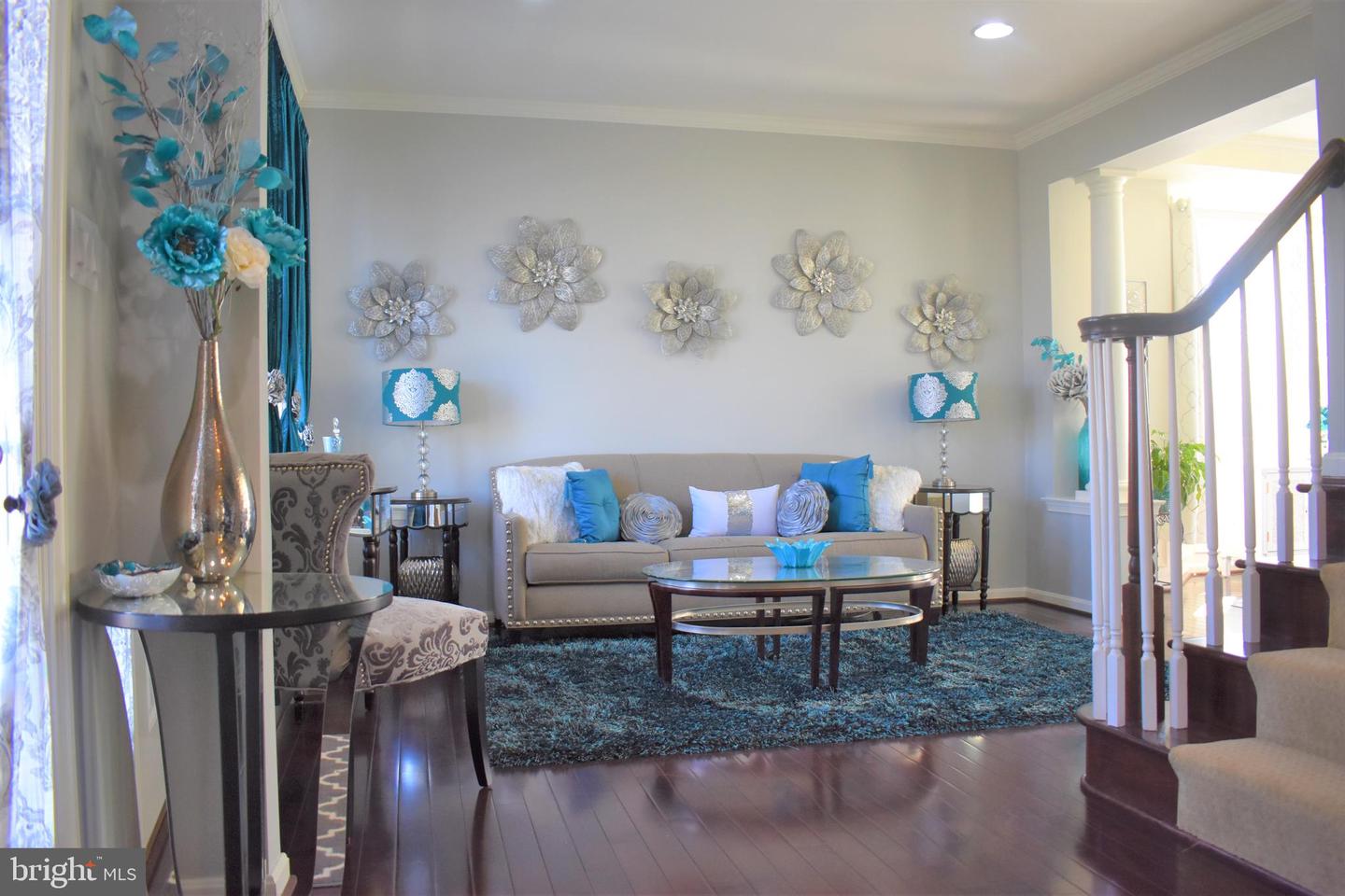 Staged living room with a beige sofa, turquoise accents, floral wall decor, and a staircase.