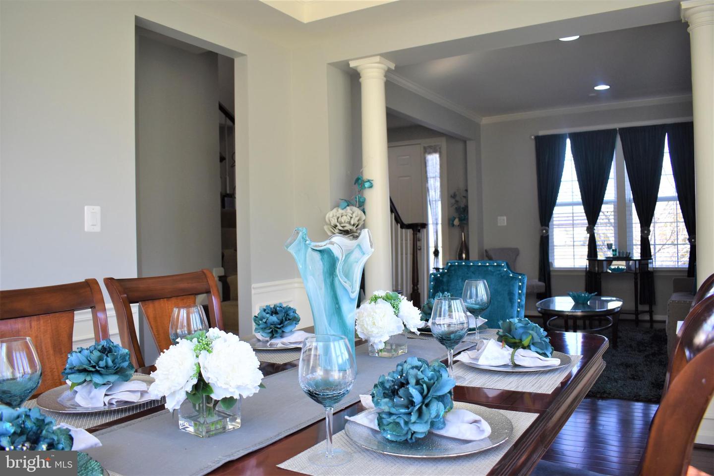 Dining room with a flower-set wooden table opening to a sitting area with teal chairs and tall curtained windows.