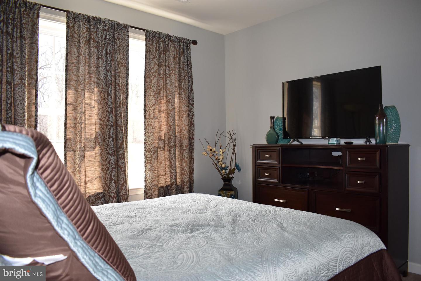 Furnished bedroom with patterned curtains, a white quilted bed, and a television on a dark dresser.
