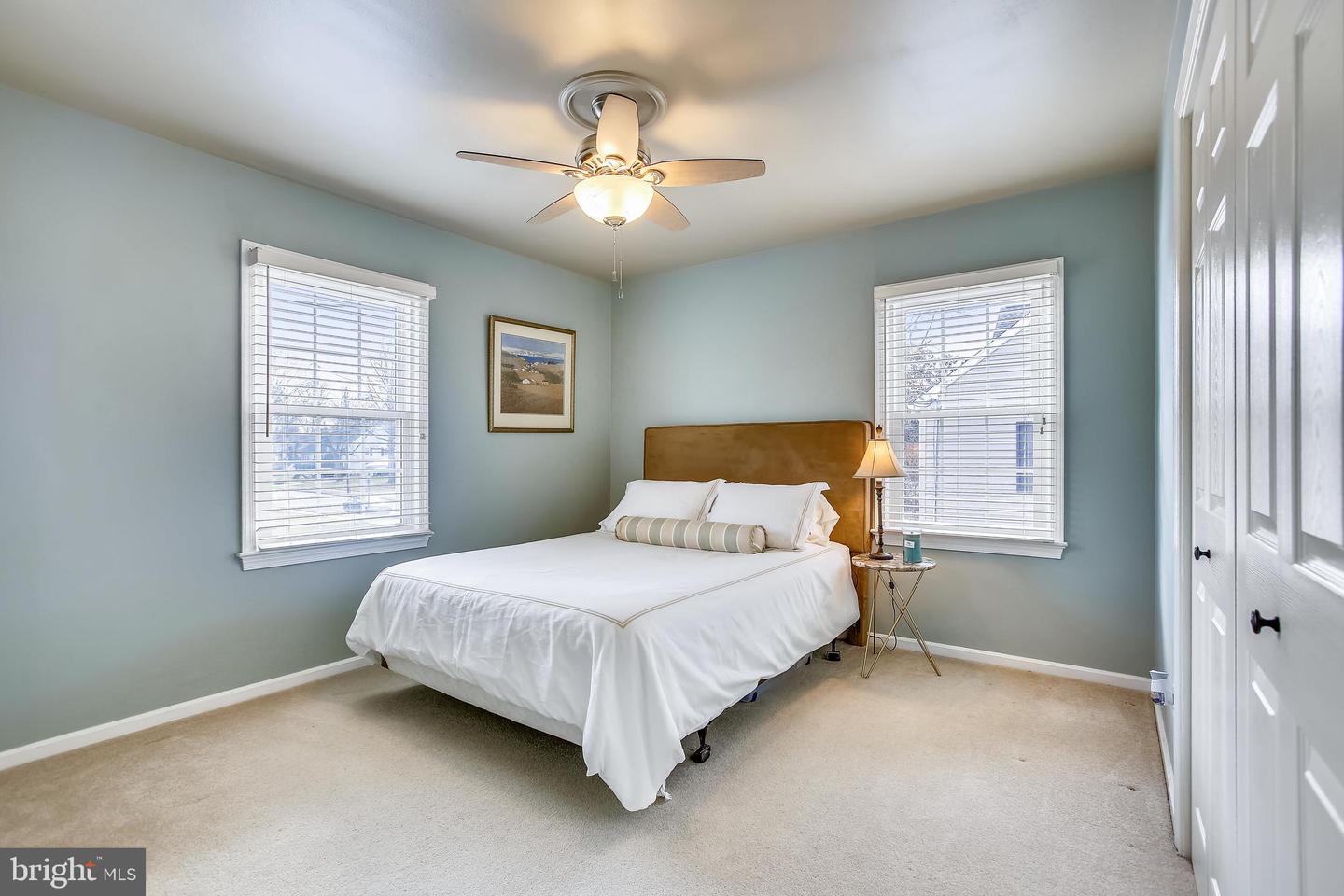 Bedroom with a white bed, tan headboard, pale blue-green walls, and two windows with blinds.