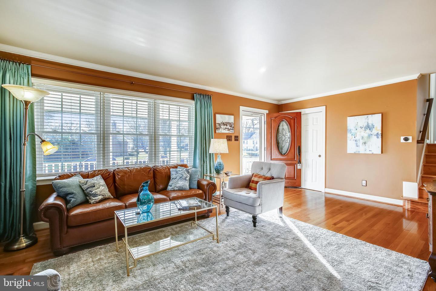 Staged living room with a leather sofa, gray armchair, and large windows overlooking the front yard.