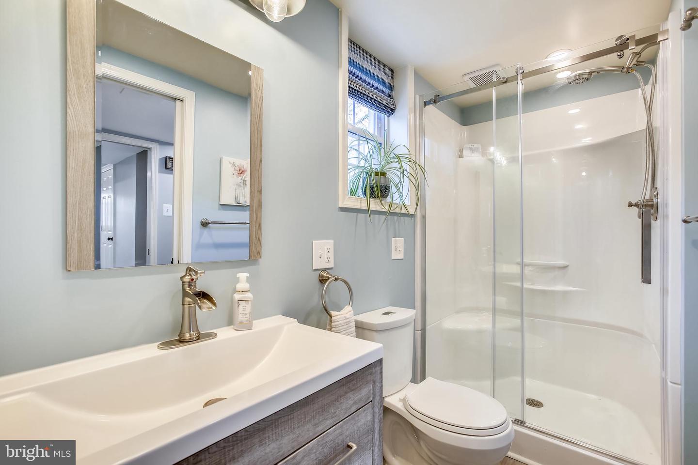 Bathroom with a wood-grain vanity, white toilet, and glass-enclosed shower beside a small window.