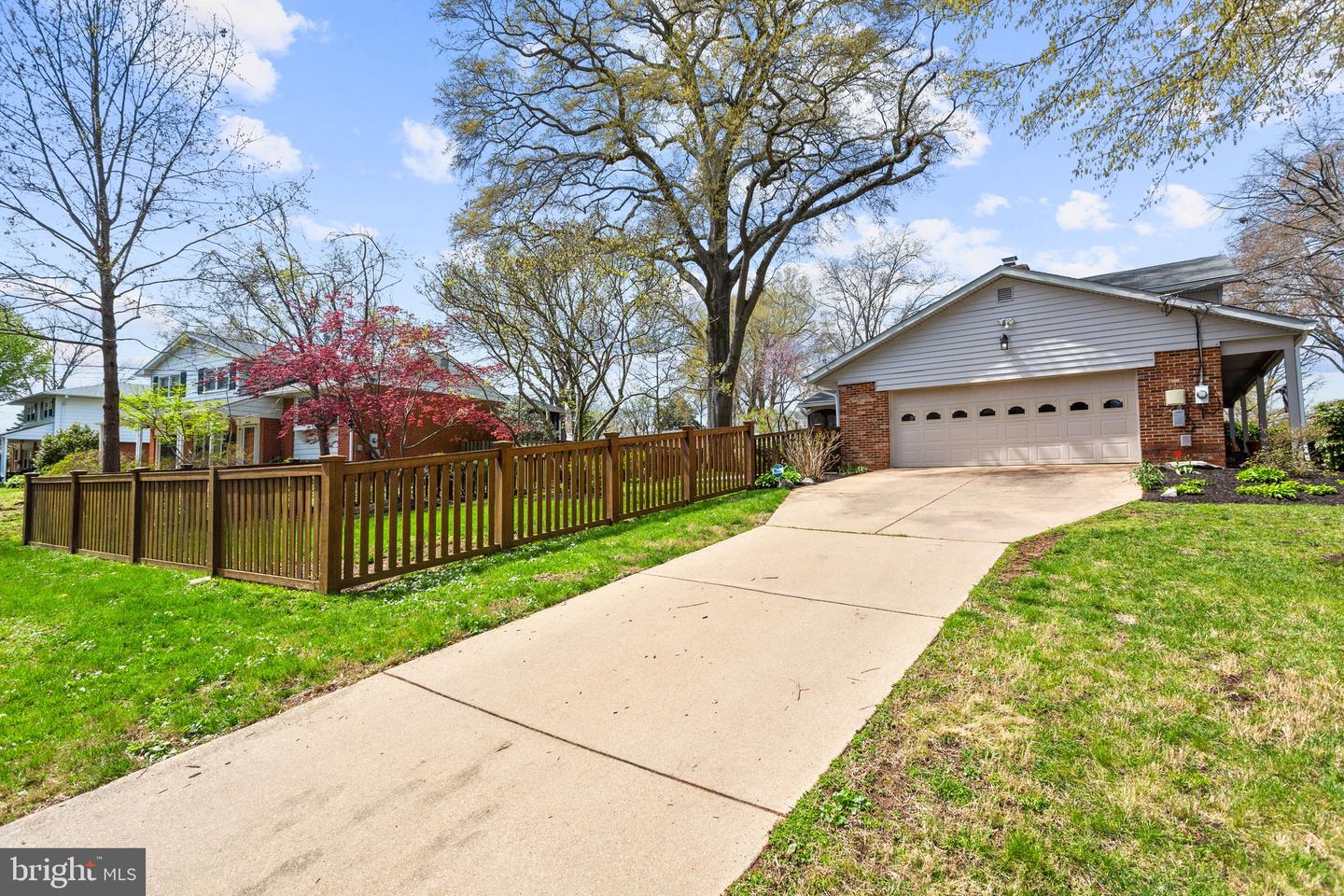 Home exterior with two-car garage, broad driveway, wooden fence, and mature trees.