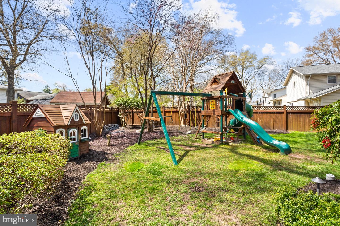 Fenced backyard with a wooden playset, green slide, swings, and small playhouse.