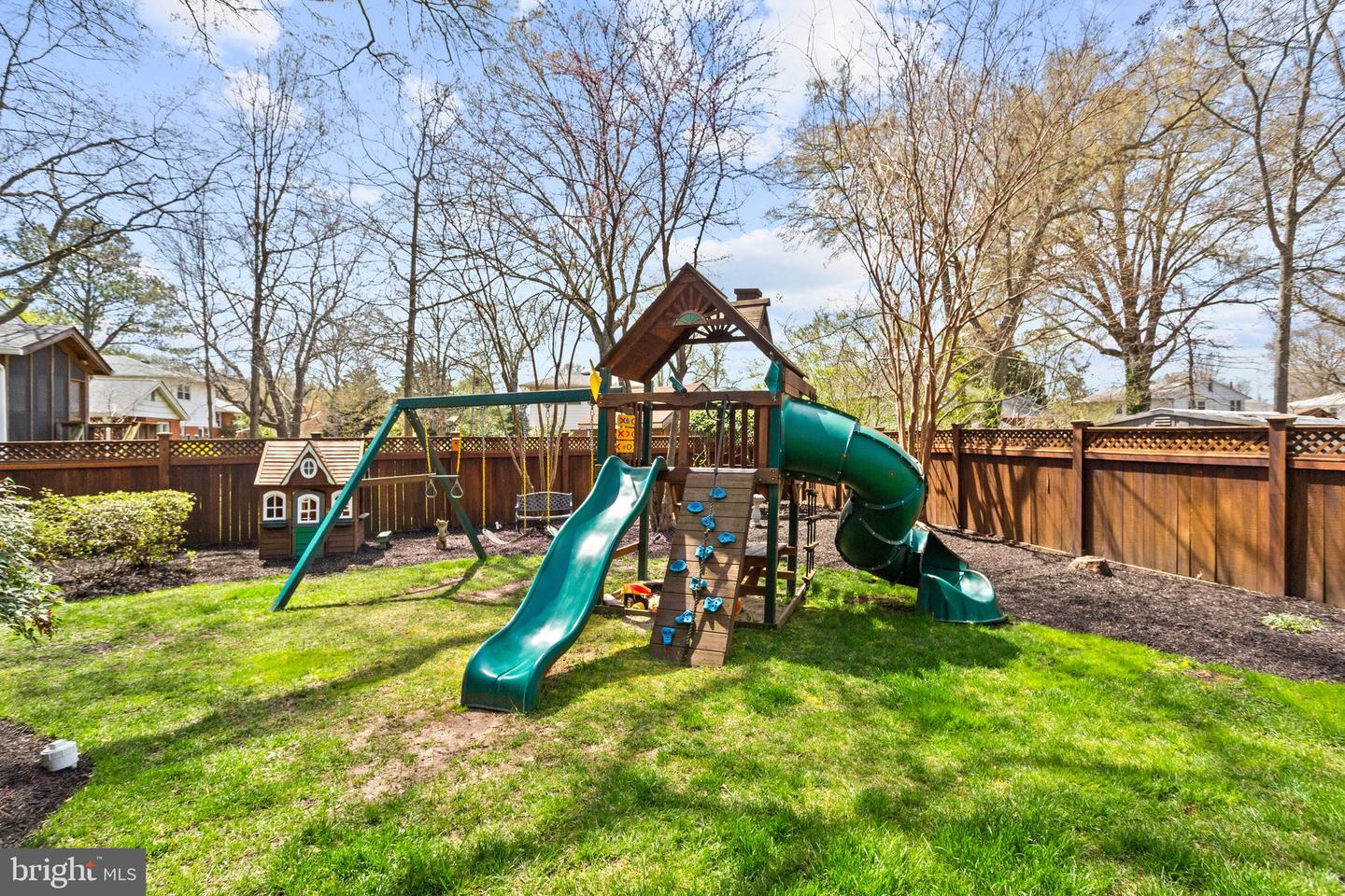 Fenced backyard with a large wooden playset, green slides, climbing wall, swings and small playhouse.