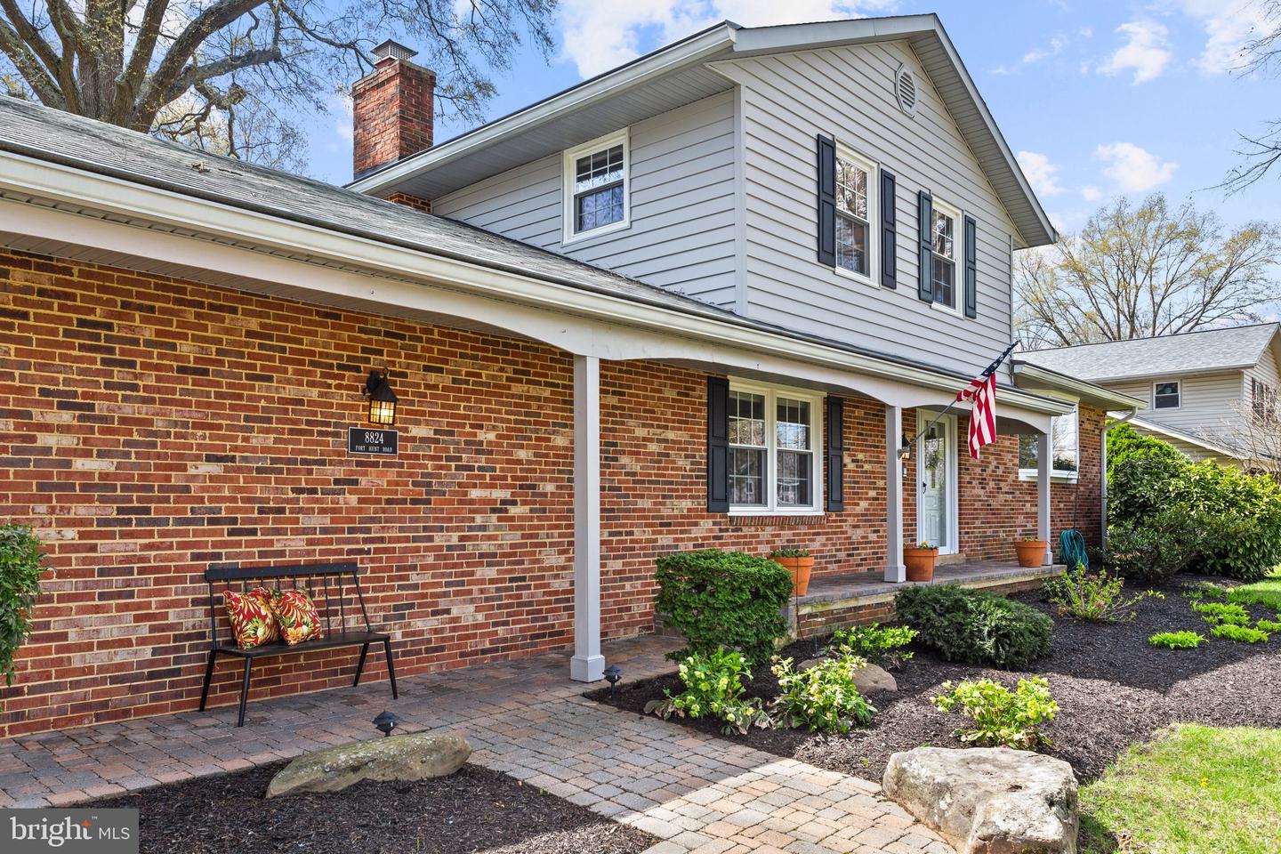 Two-story brick and white-sided home with a covered entry, landscaped beds, and an American flag.