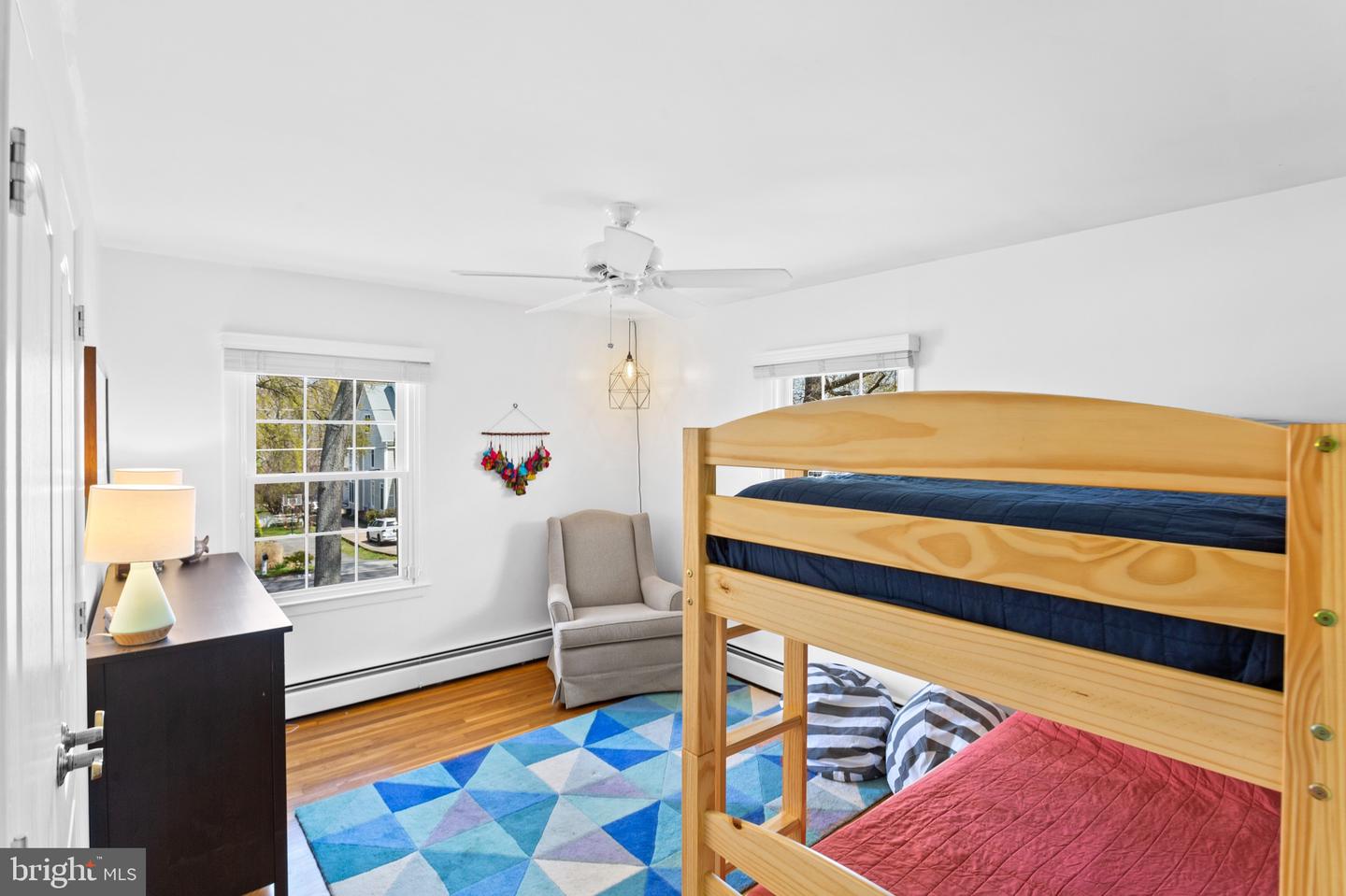 Bedroom with a wooden bunk bed, colorful geometric rug, gray armchair, and hardwood floors.