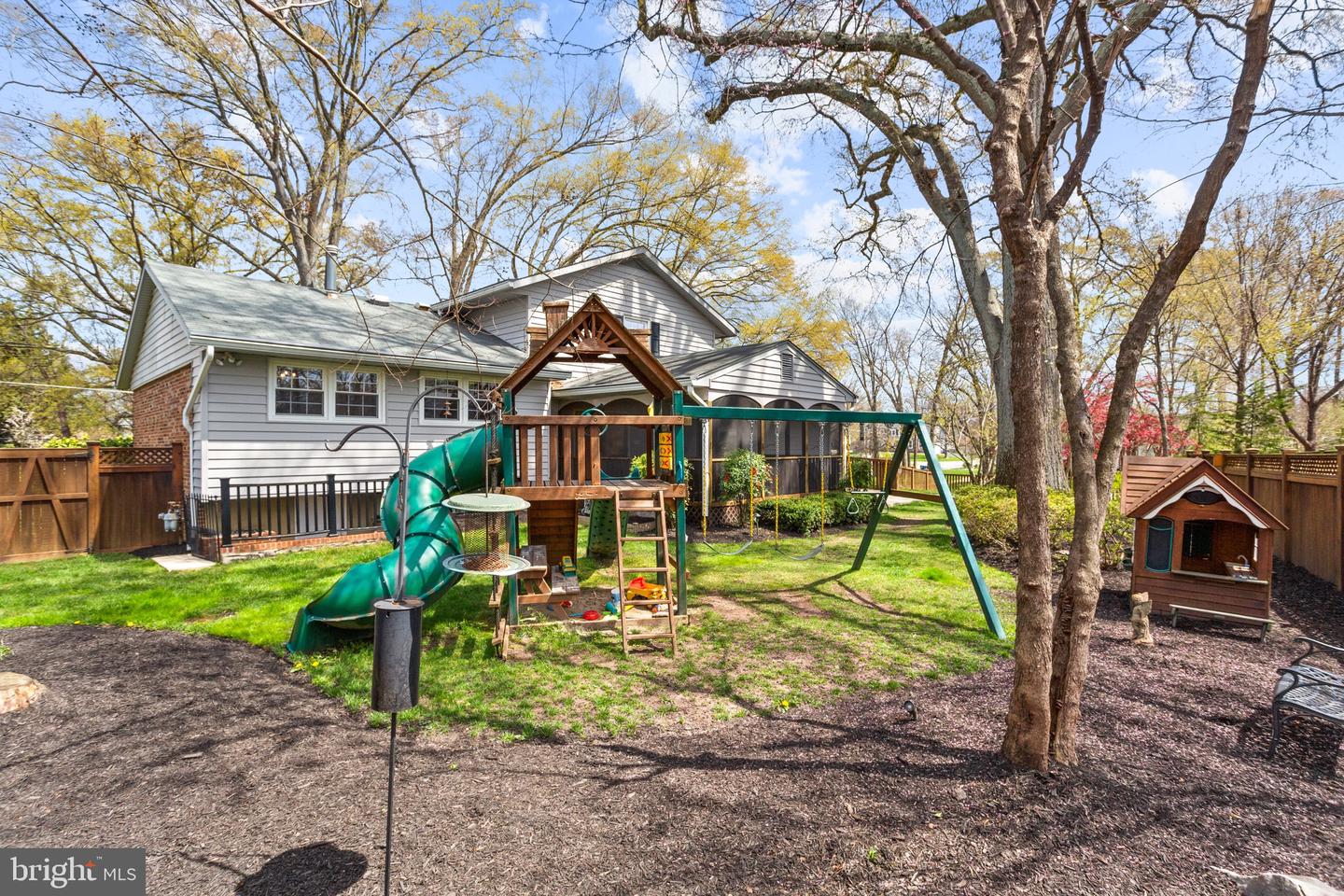 Fenced backyard with a large playset, swings, green slide, small playhouse, and mature trees.
