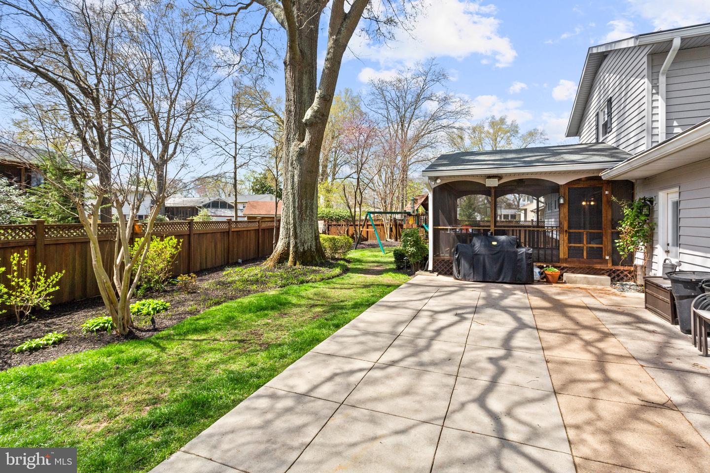 Backyard patio beside a screened porch, with a fenced lawn, mature tree, and swing set.
