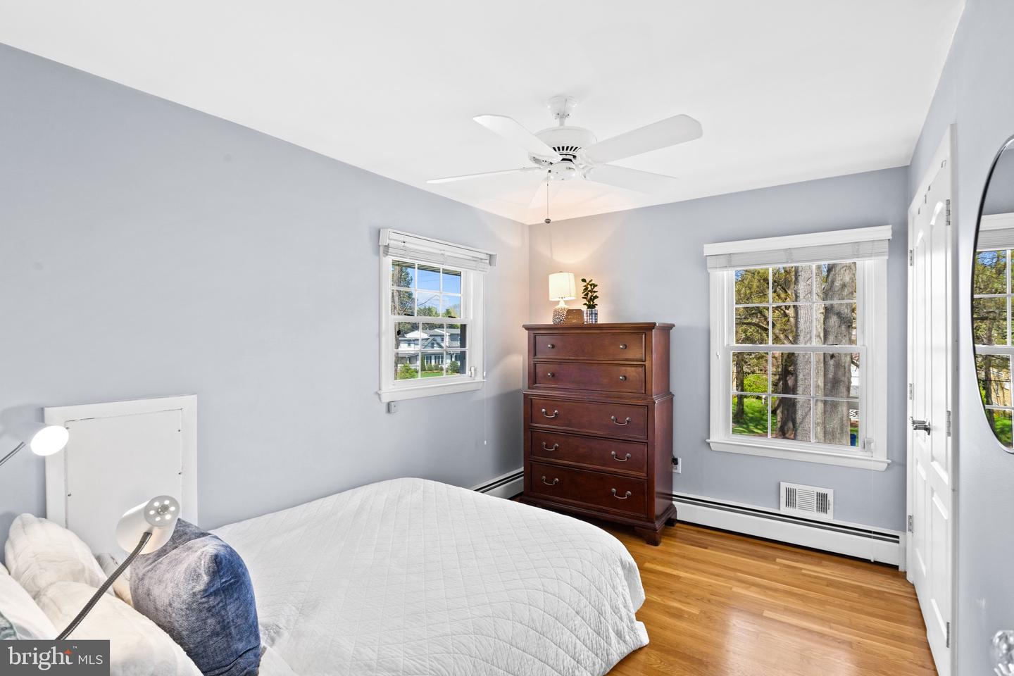 Bedroom with a white bed, dark wood dresser, ceiling fan, and two windows overlooking trees.