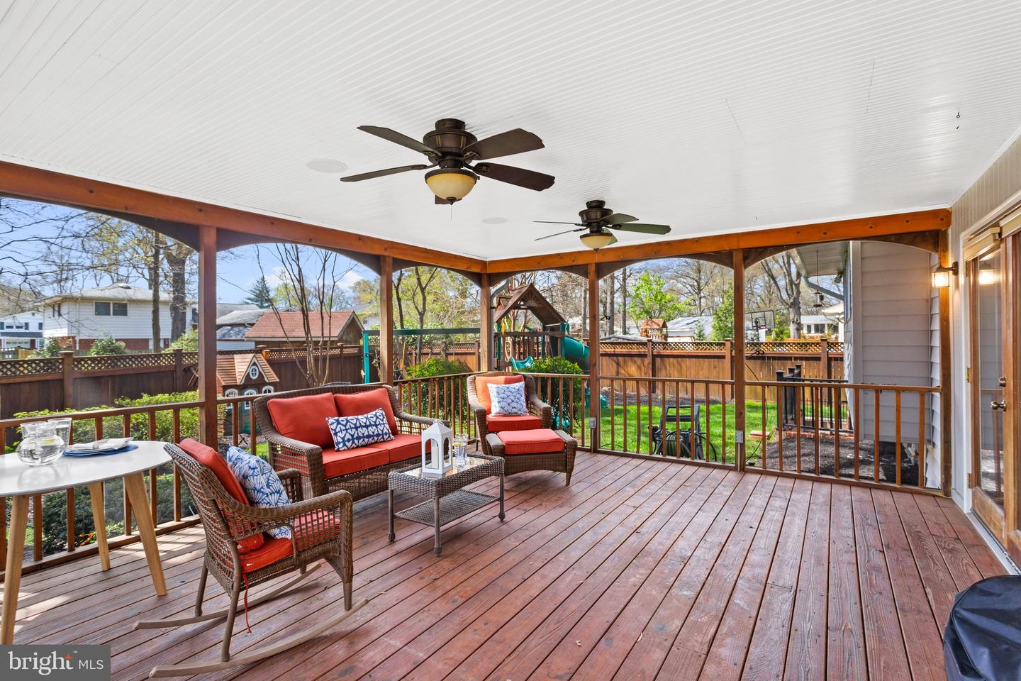 Covered wooden deck with wicker seating and red cushions overlooking a fenced backyard and playset.