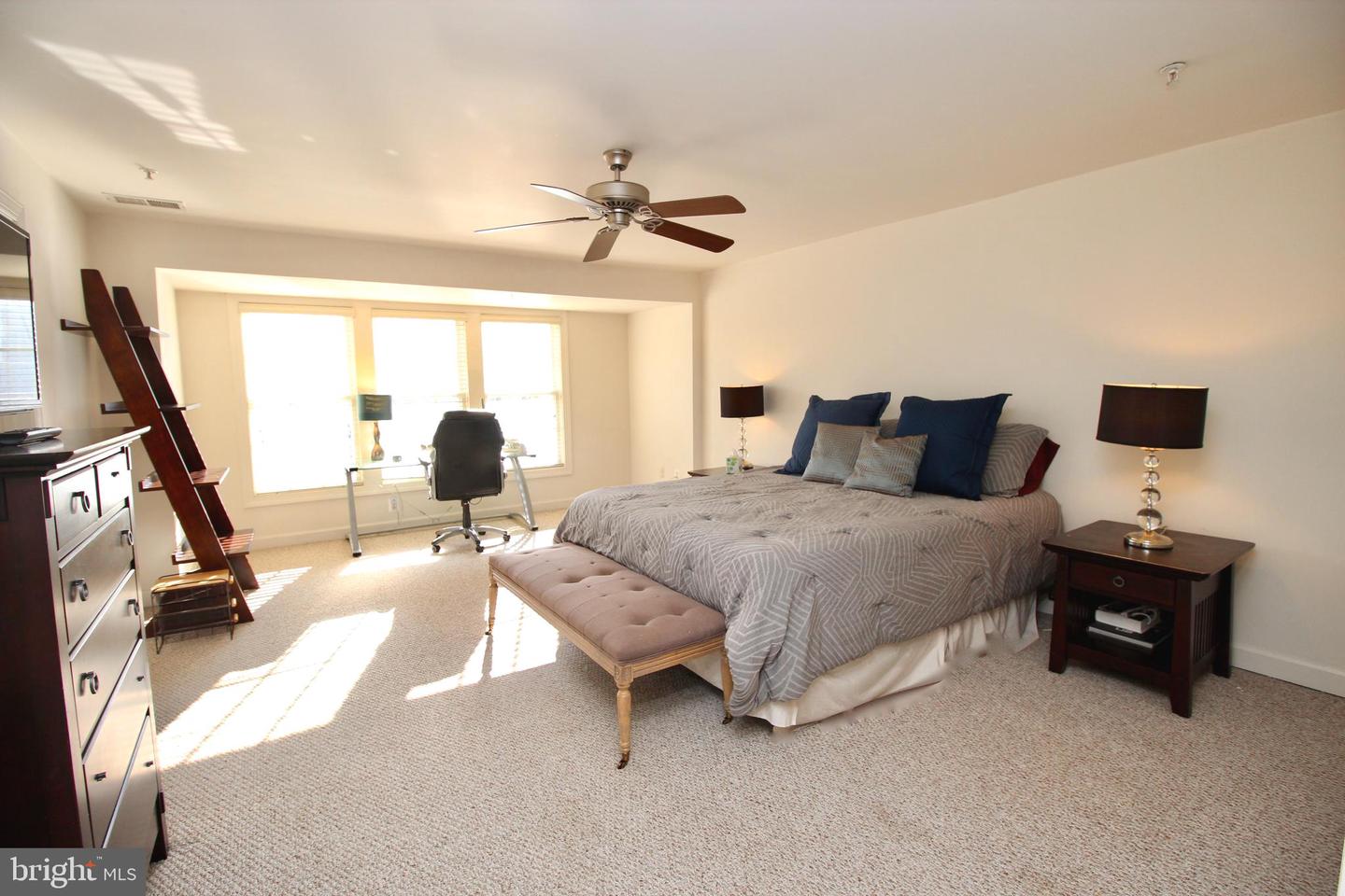 Furnished bedroom with a large gray-covered bed, navy pillows, tufted bench, and sunny windows.