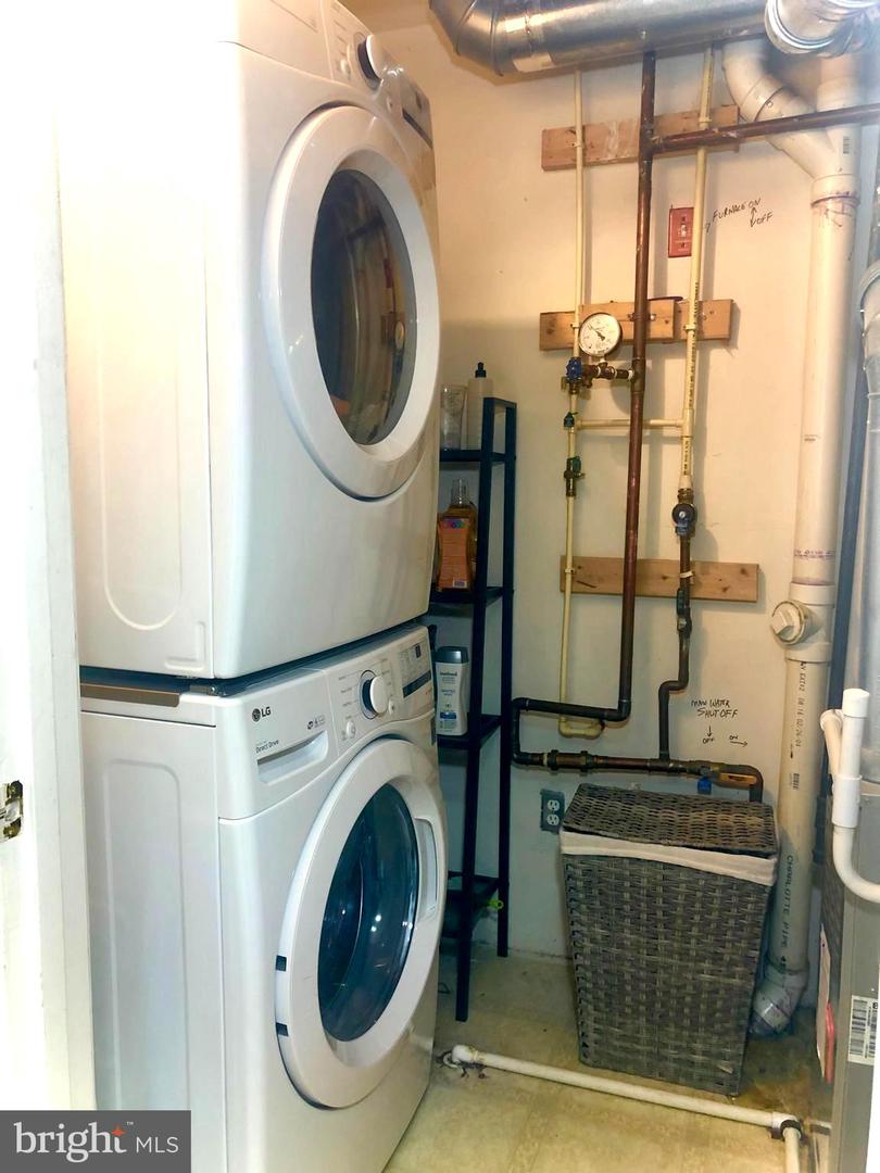 Laundry room with stacked front-loading washer and dryer, exposed utility pipes, and a woven hamper.