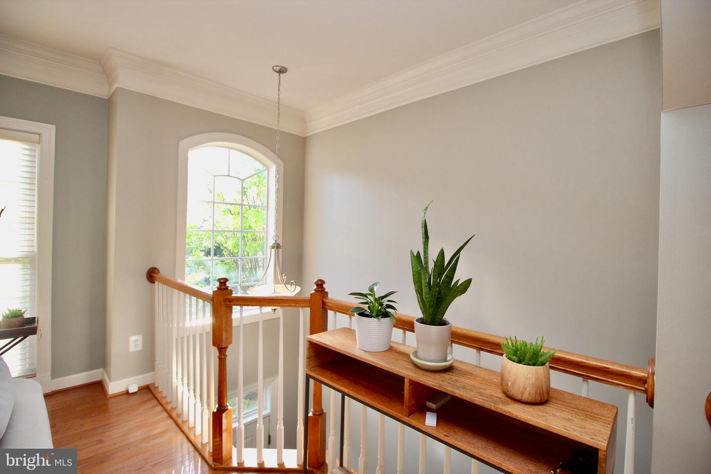 Upstairs landing with a polished wood stair railing, arched window, and console table with potted plants.