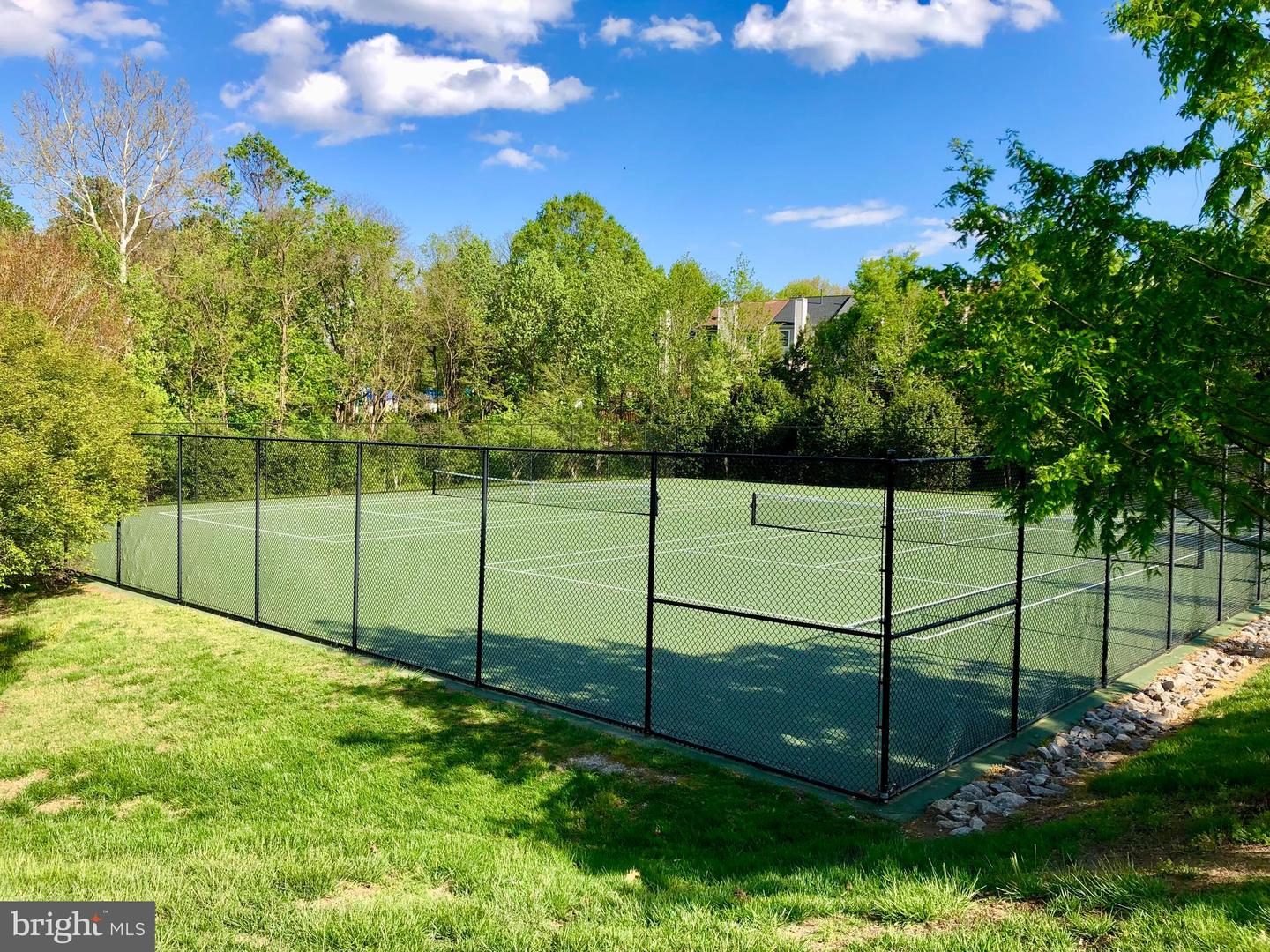 Outdoor tennis court enclosed by black fencing, surrounded by trees and lawn.