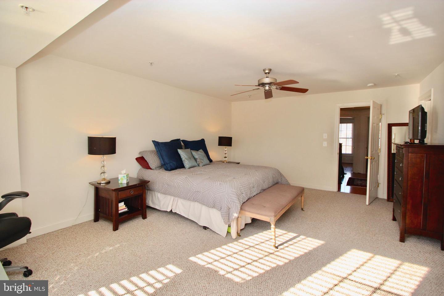 Furnished bedroom with a large bed, cushioned bench, ceiling fan, and sunlight across the carpet.
