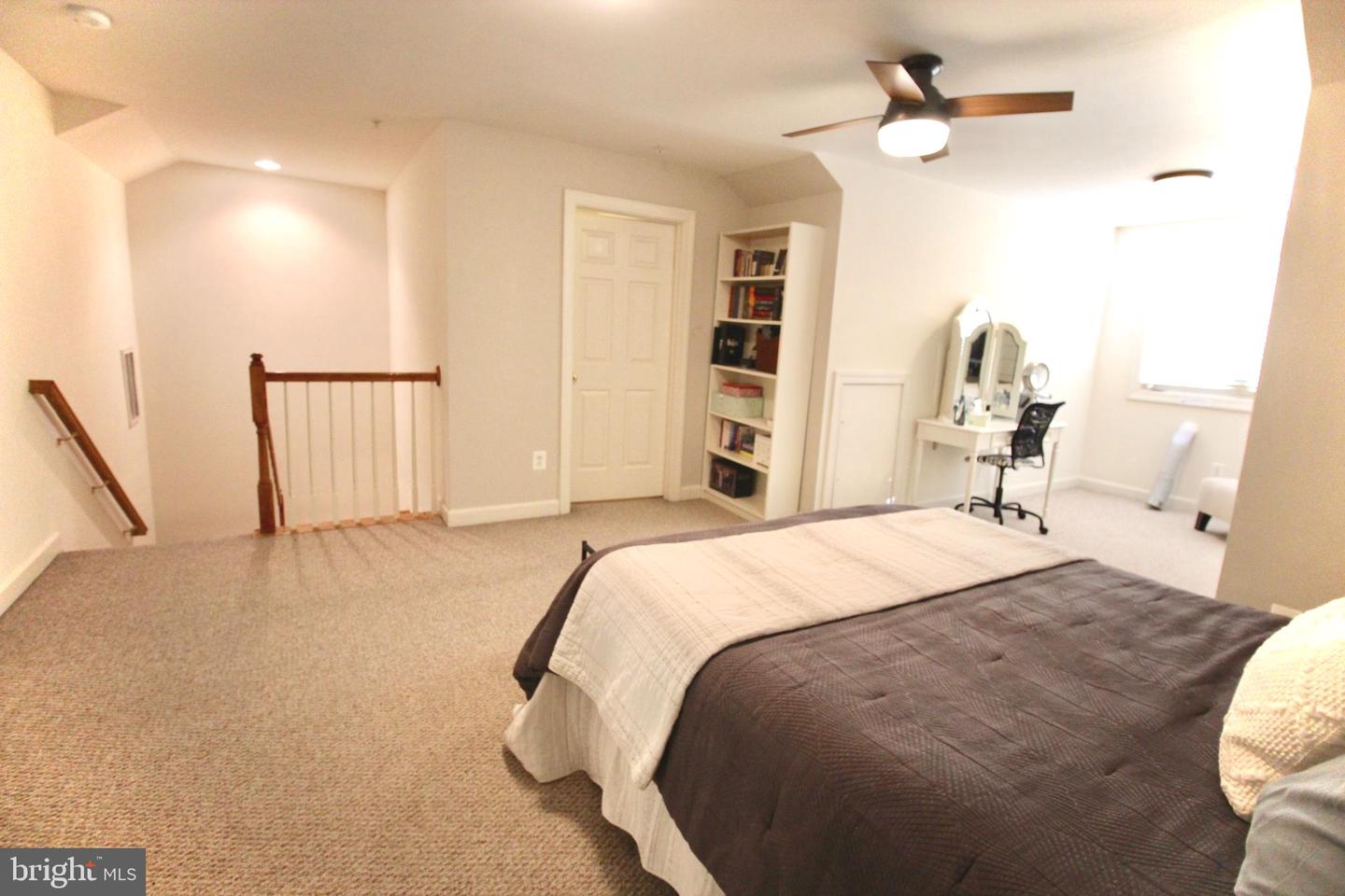 Furnished bedroom with a bed, ceiling fan, desk, and open stair railing.