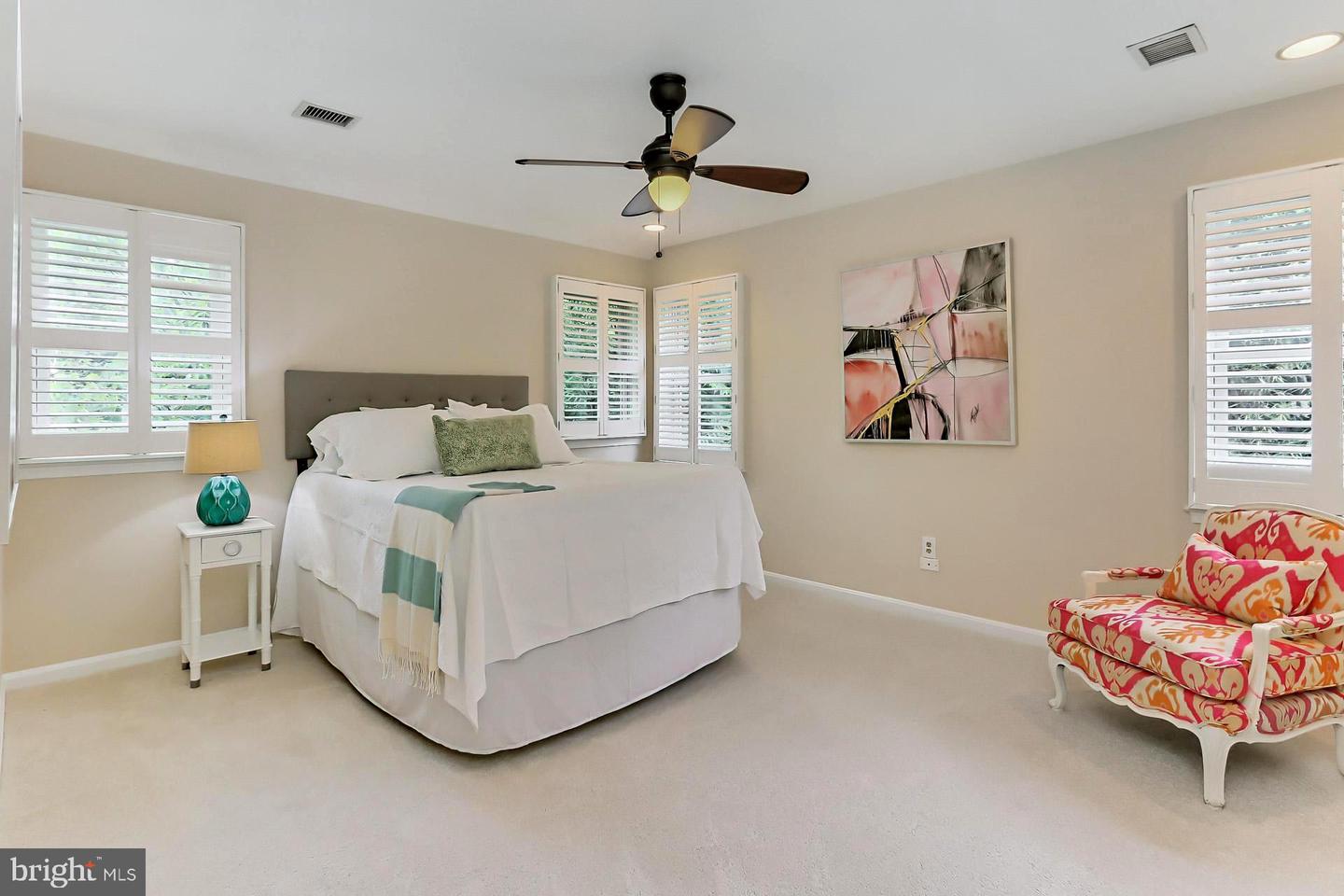 Staged bedroom with a white bed, plantation shutters, ceiling fan, and colorful armchair.
