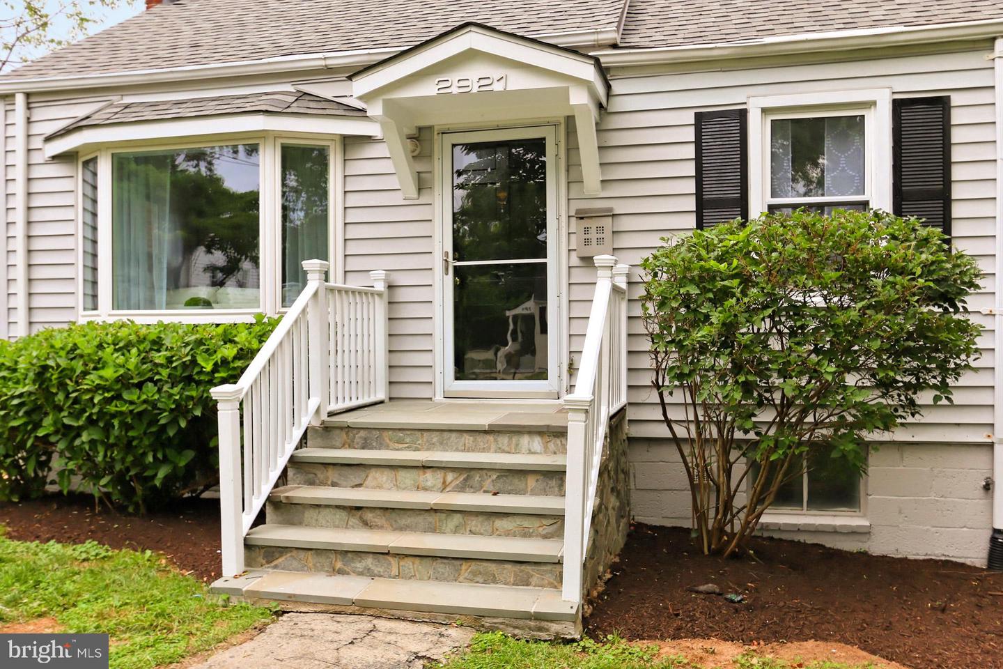 House front with covered entry, stone steps, white railings, bay window, and shrubs.