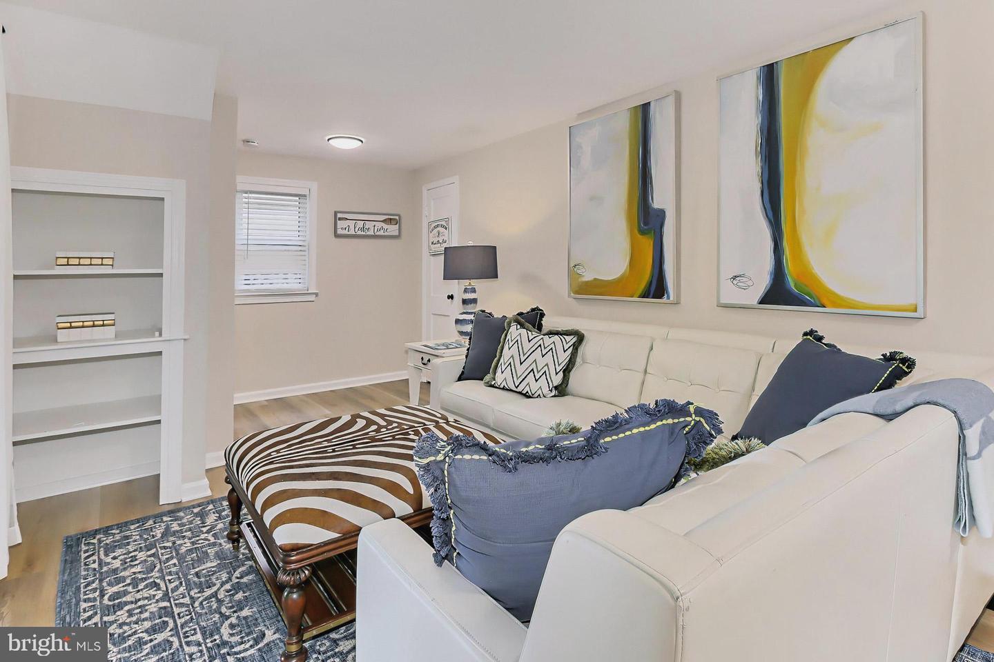 Staged living room with white sofas, a zebra-striped ottoman, built-in shelves, and colorful abstract art.