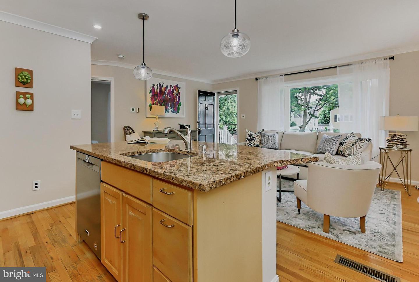 Open-plan kitchen with a granite island and wood cabinets beside a bright, furnished living area.