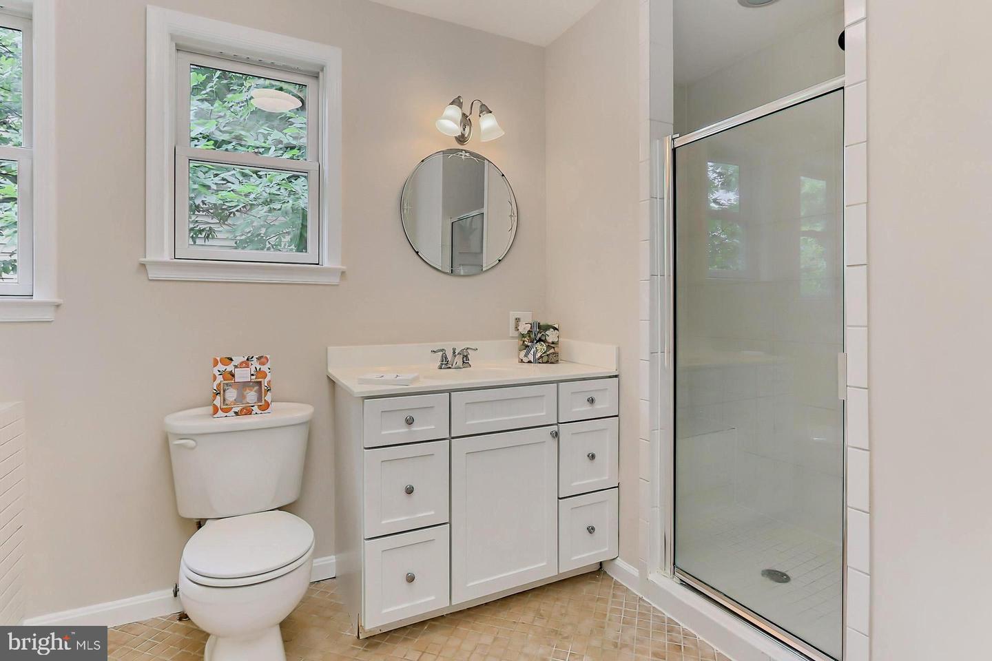 Bathroom with a white vanity, round mirror, and glass-enclosed shower beside two windows.