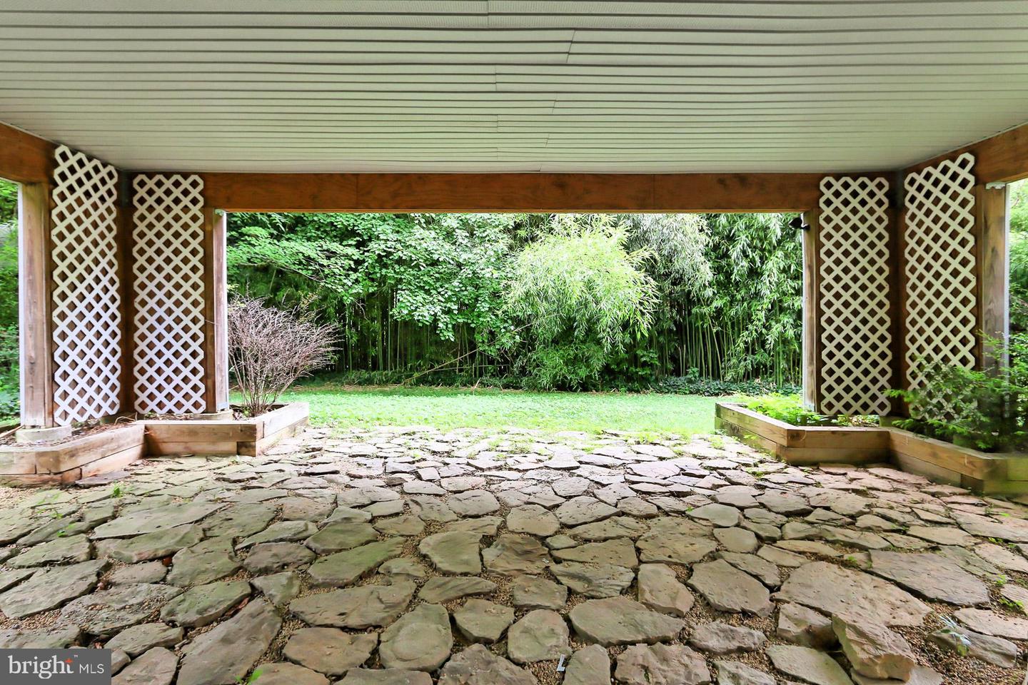 Covered stone patio with white lattice panels, raised planters, and a green lawn backed by trees.