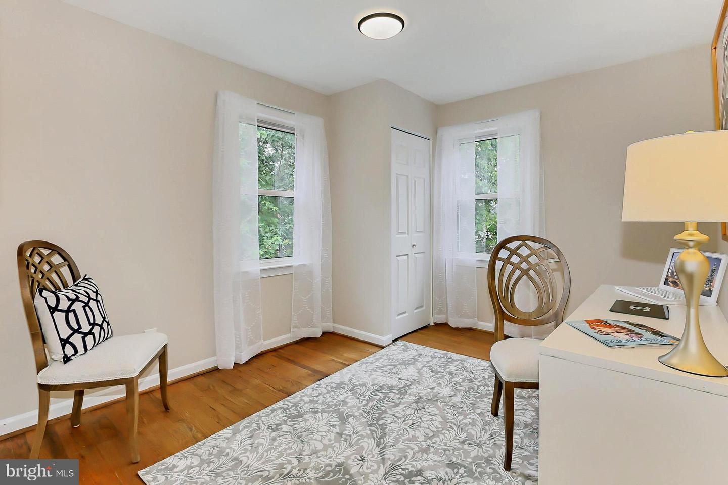 Staged bedroom with a patterned bedspread, hardwood floors, and two curtained windows.