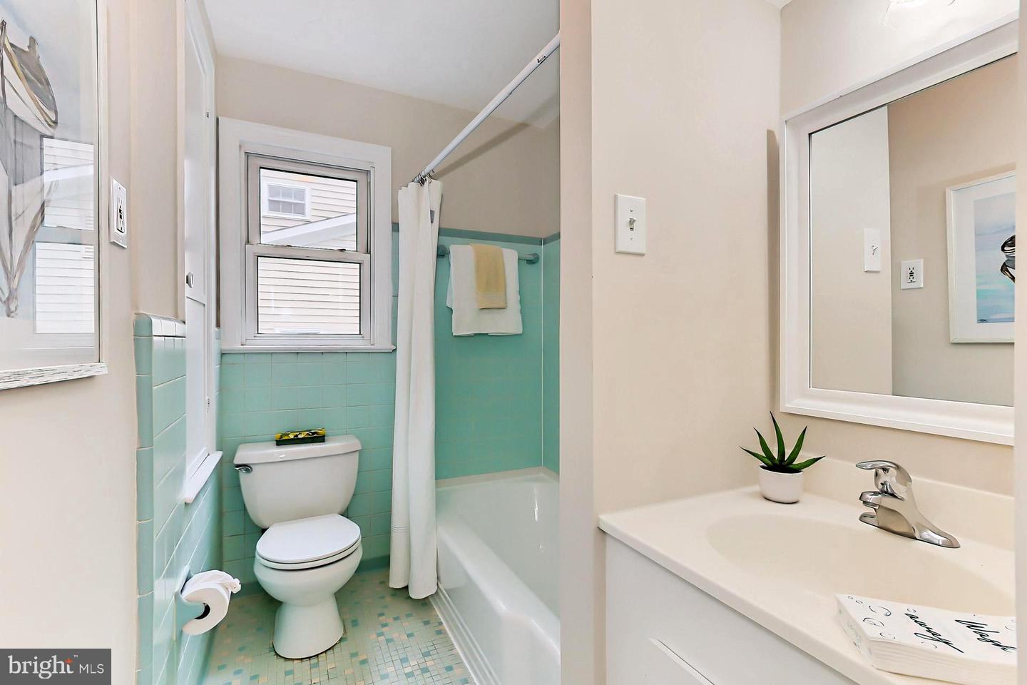 Bathroom with turquoise tile, a tub-shower, toilet beneath a window, and white vanity with mirror.