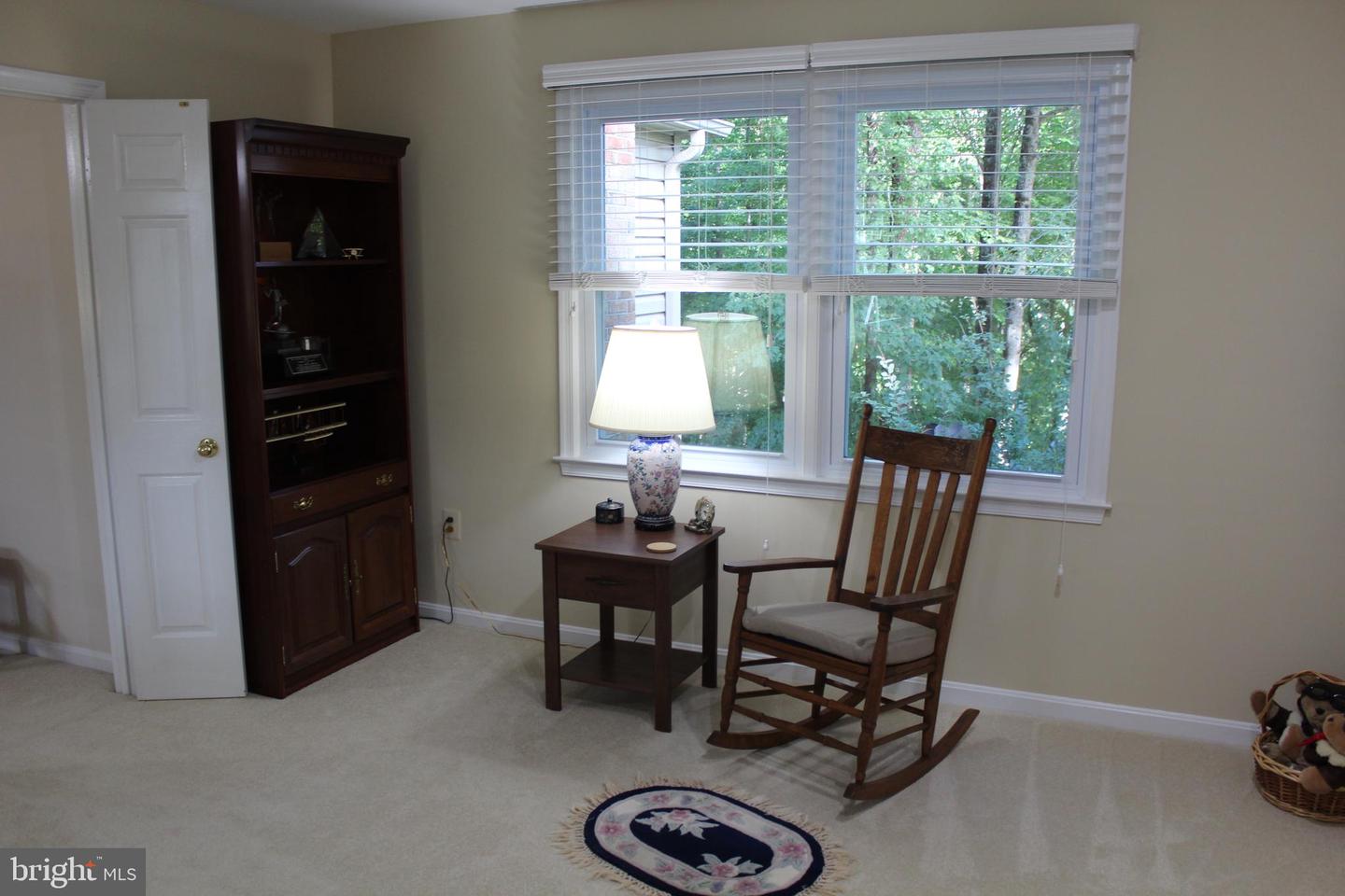 Sitting area with a wooden rocking chair and lamp beside a large window overlooking trees.
