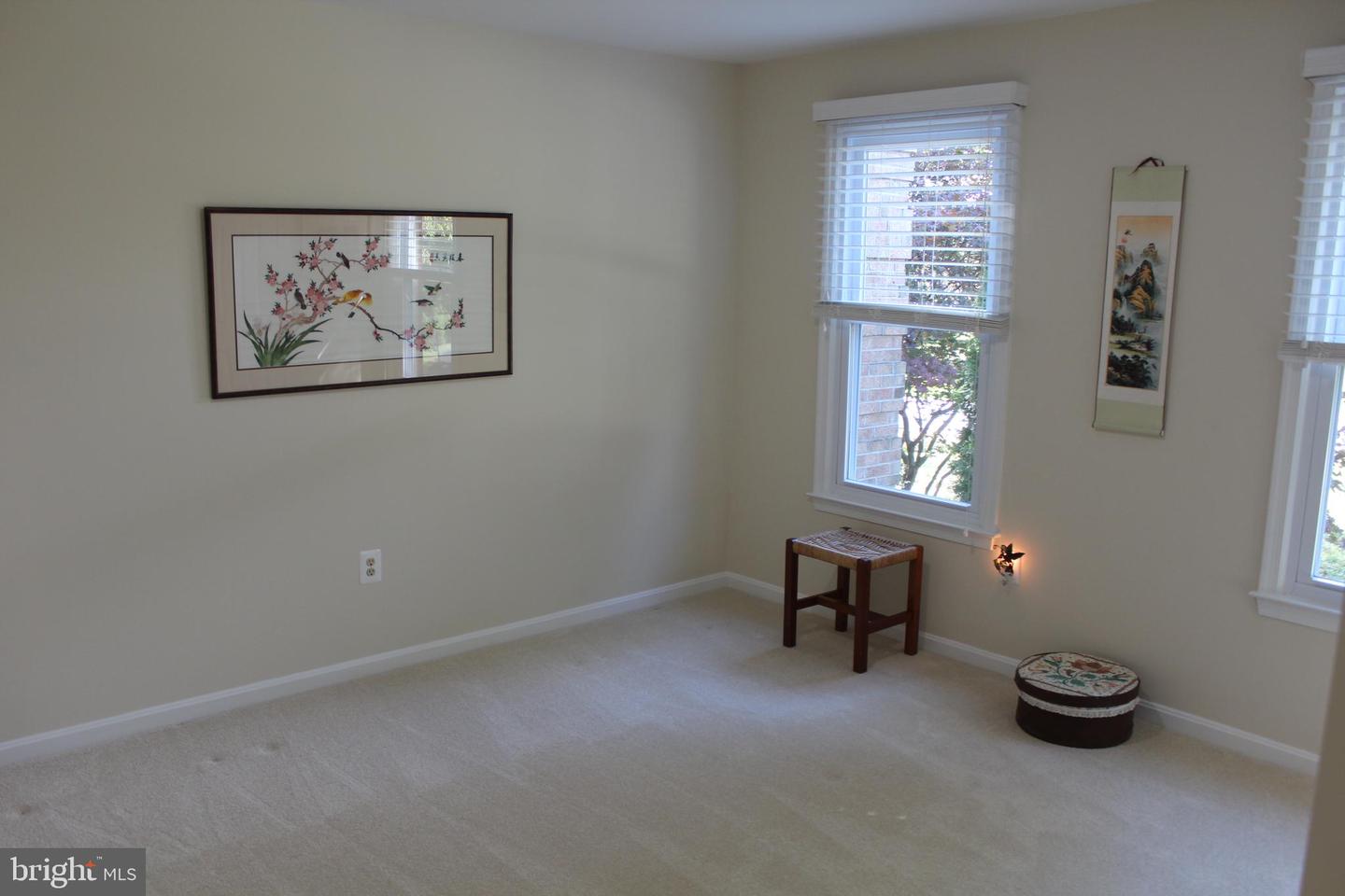 Carpeted room with two windows, framed artwork, a wooden stool, and a round ottoman.