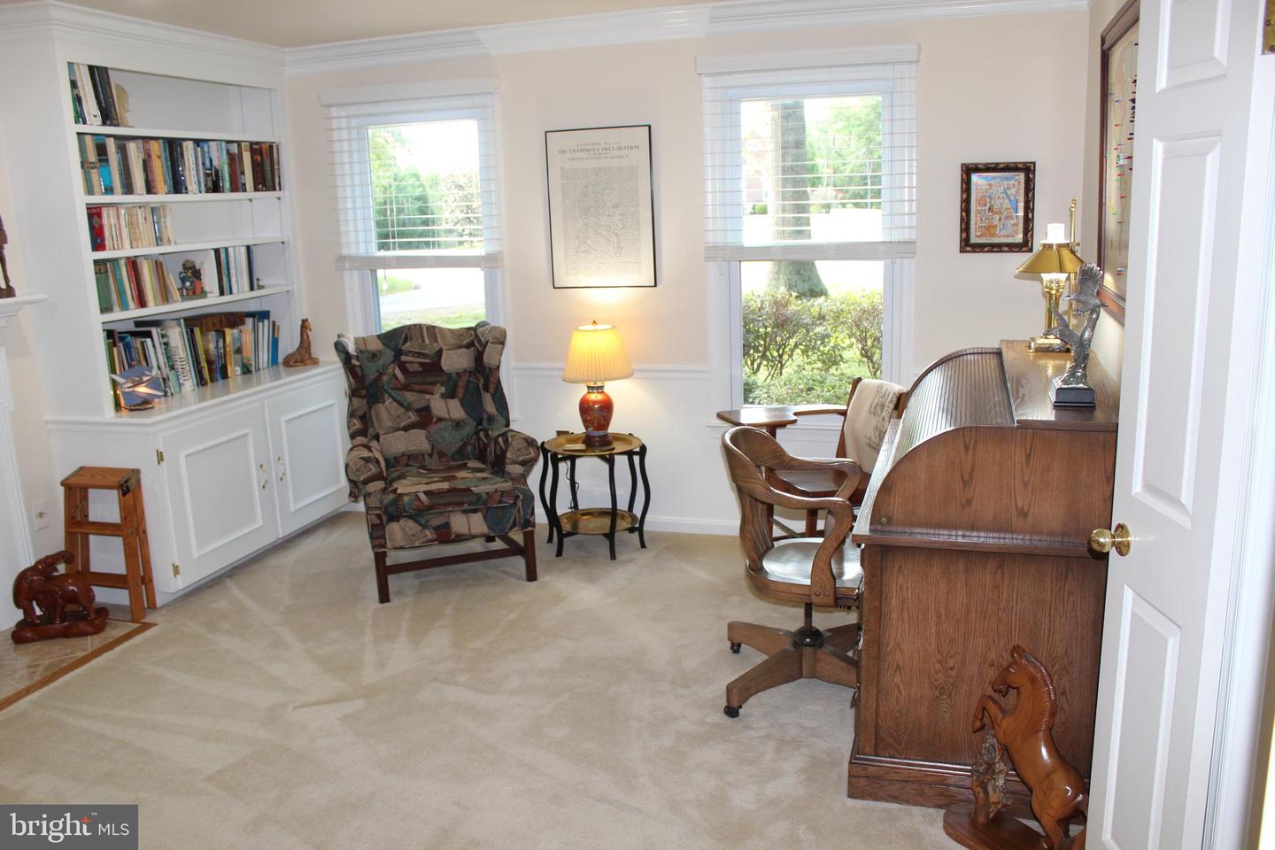 Furnished home office with built-in bookshelves, a wooden roll-top desk, and two bright windows.
