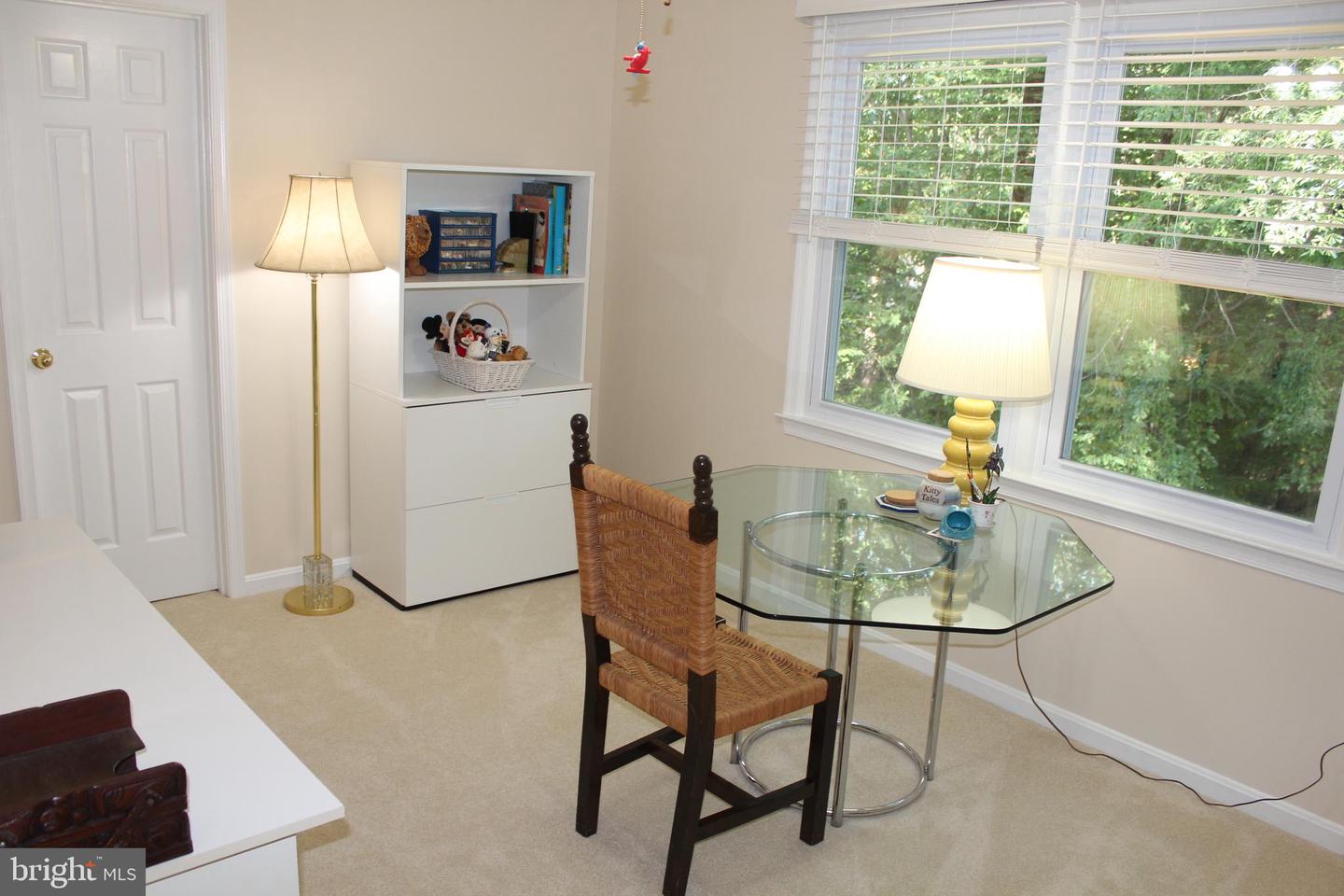 Furnished room with a glass-top table and wicker chair beside large windows overlooking greenery.