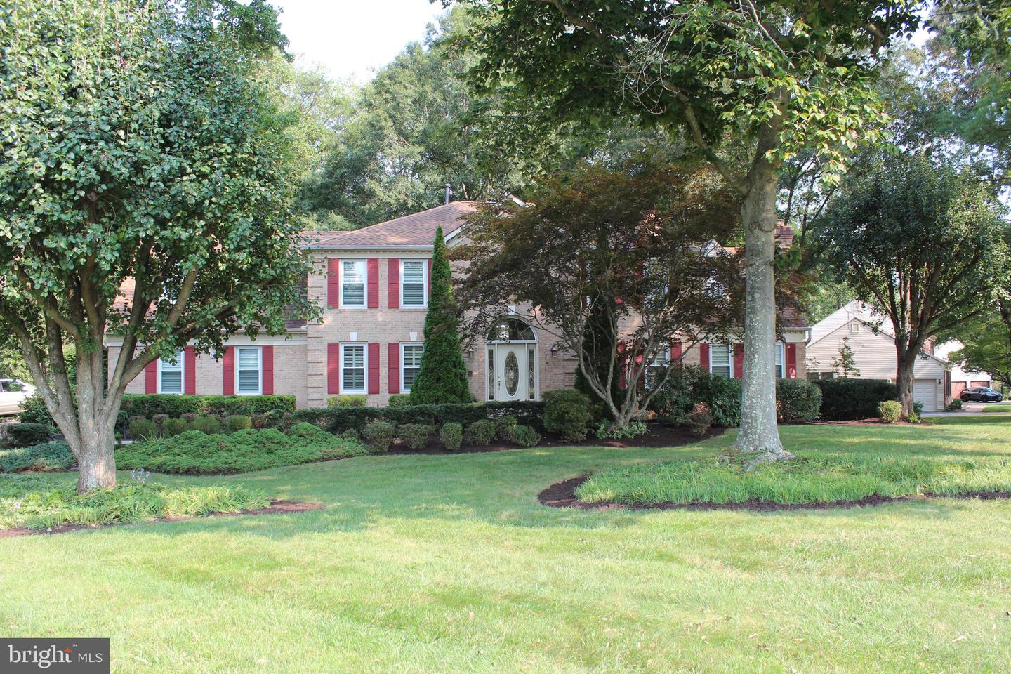 Two-story brick house with red shutters, mature trees, and a landscaped front lawn.