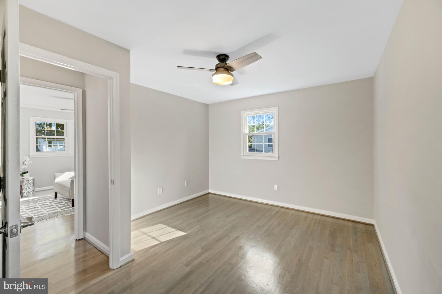 Empty bedroom with light wood flooring, a ceiling fan and a window; doorway opens to a furnished room.